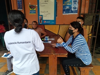 A woman in a white shirt with the words "humanitarian assistance" speaks with a small group of migrant women and men who are seated around a wooden table. 