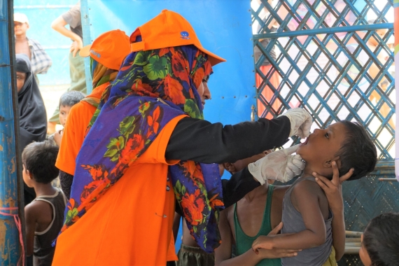 A female health care worker in orange jacket and hat cares for a young child at an outdoor health screening in Bangladesh.
